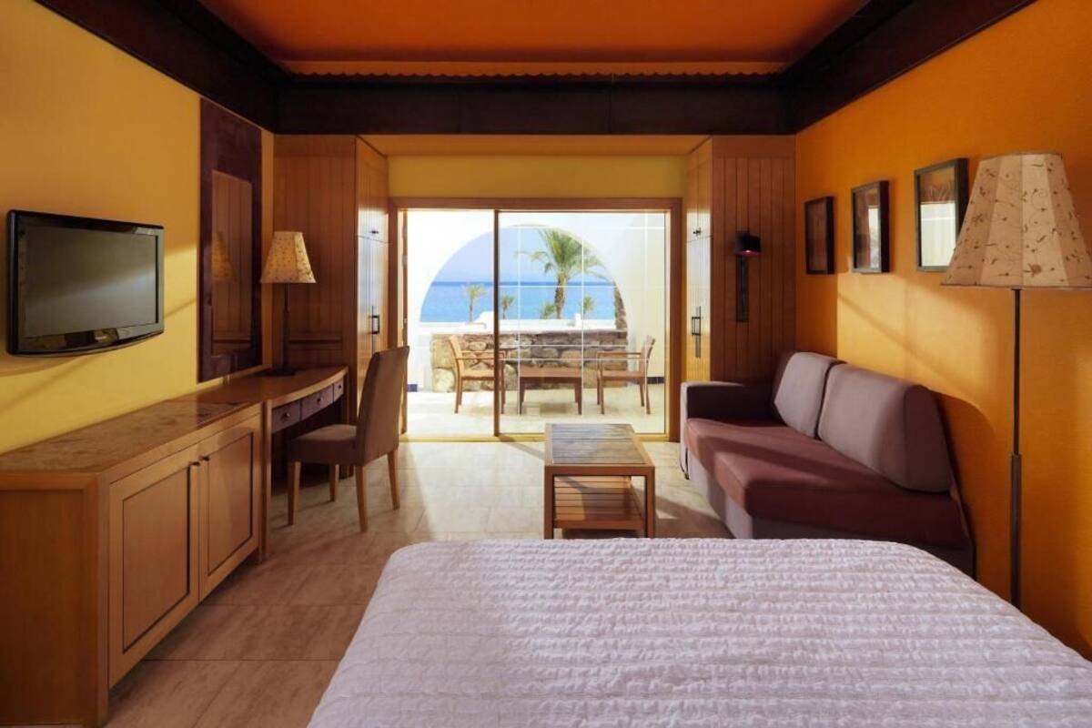 Ett hotellrum med en säng, ett skrivbord, en tv, en soffa och utsikt över havet.