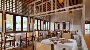Un amplio comedor con varias mesas y sillas, ventanas grandes que ofrecen vistas a palmeras y una estructura de techo de madera.