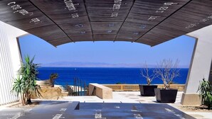 Un espacio moderno al aire libre con vista al mar, que presenta un suelo de hormigón con texto, plantas en macetas y una cerca de madera.