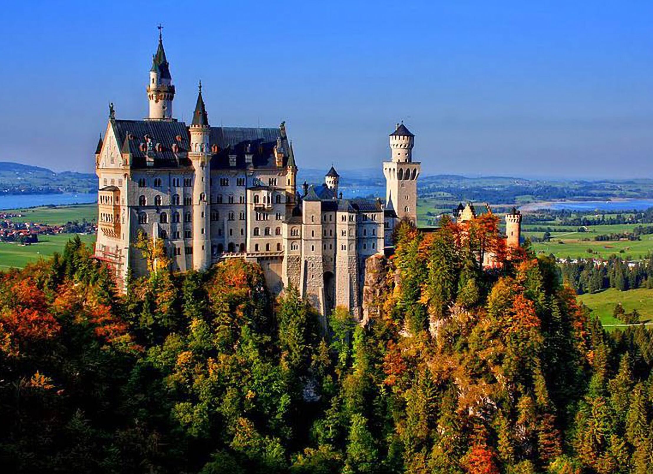 A historic castle with multiple towers and a prominent central building, surrounded by autumn foliage and a scenic landscape.