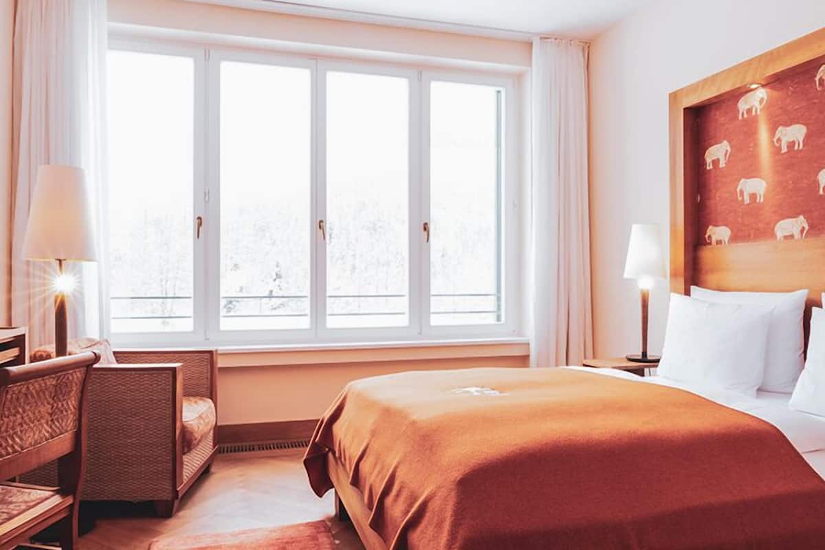 A hotel room with a large window, a bed with a brown cover, a chair, a nightstand with a lamp, and a wall with a wooden panel featuring animal silhouettes.