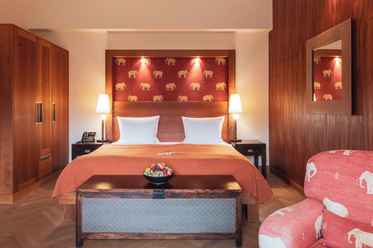 A hotel room with a large bed, wooden headboard, two bedside tables with lamps, a wooden wardrobe, and a red patterned sofa.