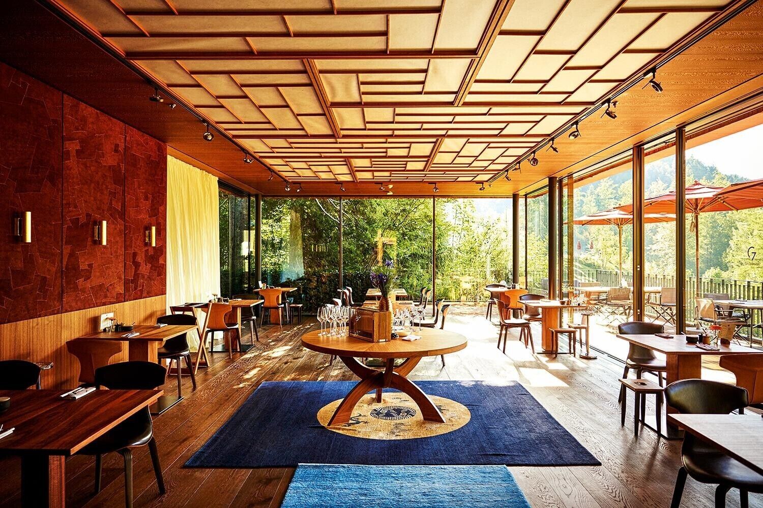 A modern restaurant with a wooden ceiling, large windows, and a central round table.