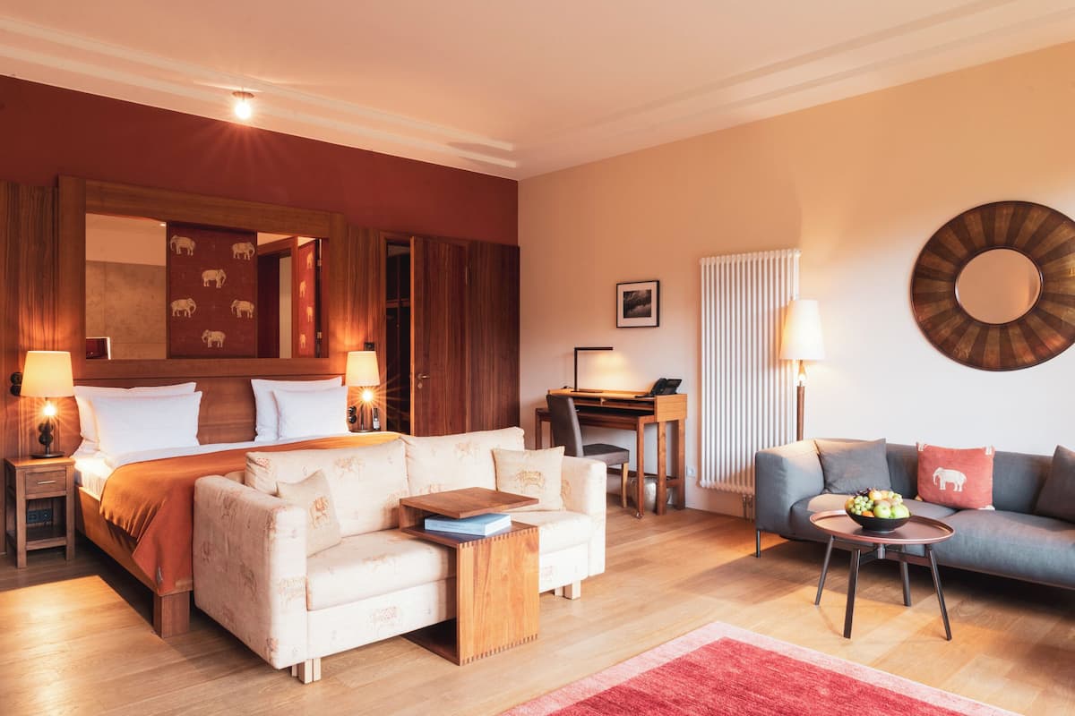 A well-lit hotel room with a large bed, a sofa, a desk, and a dining area.