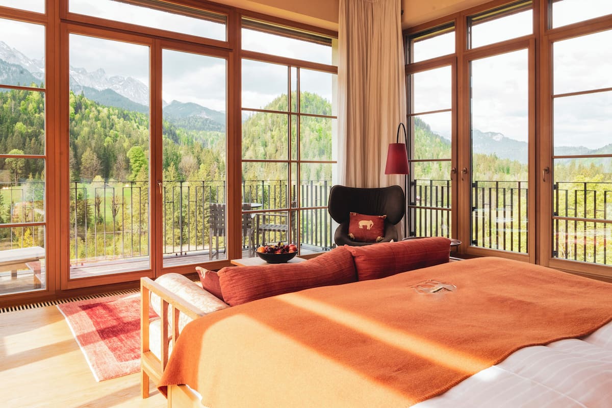 A bedroom with a large bed, a view of a forest and mountains through large windows, and a balcony with a railing.