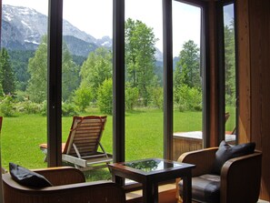 A room with large windows offering a view of a green landscape and mountains. There is a wooden chair and a small table with a glass top. - Schloss Elmau Luxury Spa Retreat & Cultural Hideaway (Kruen)