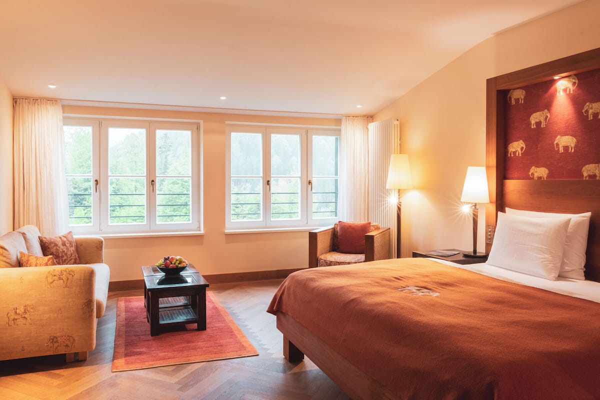 A hotel room with a bed, a sofa, a small table with fruit, and a view of greenery through large windows.