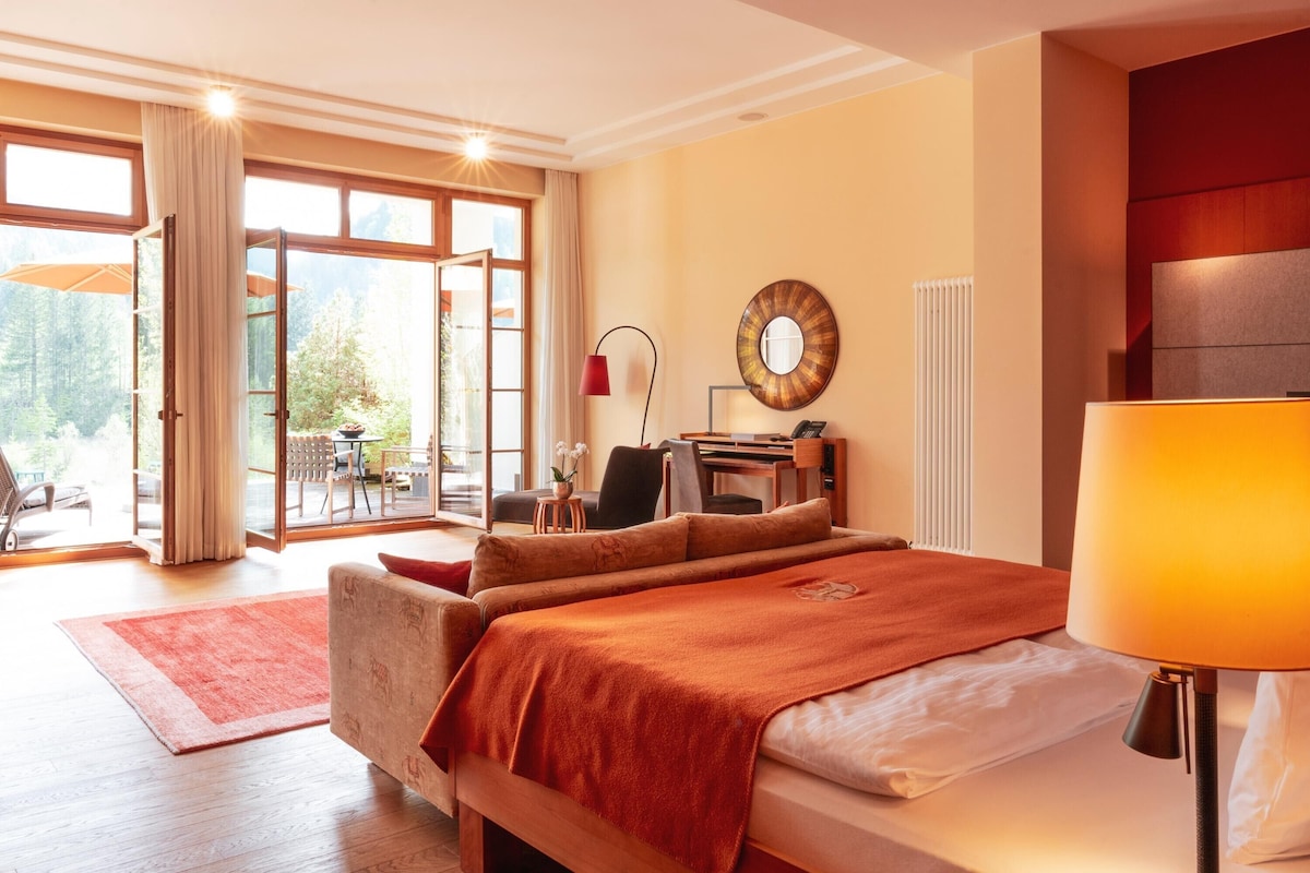 A hotel room with a bed, a desk, a chair, a lamp, and a view of the outdoors through large windows.