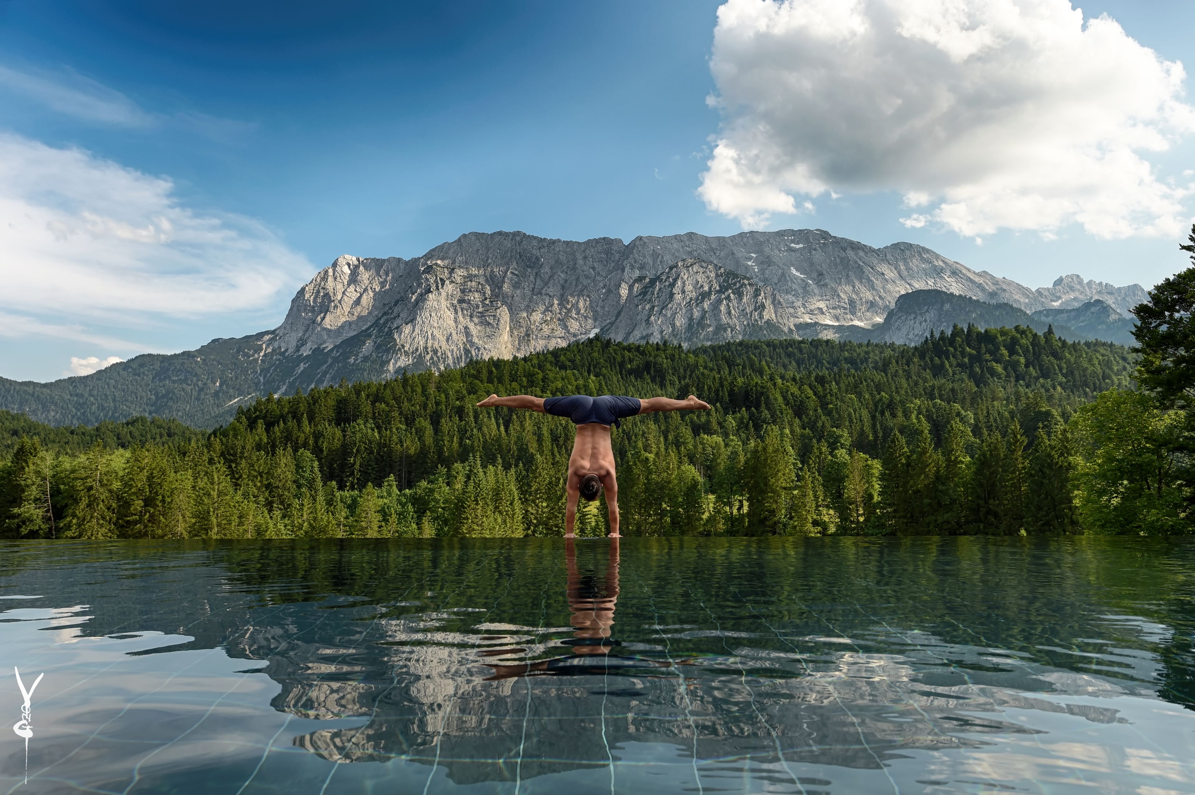 Una persona haciendo una postura de parada de manos en una piscina cristalina y reflectante, con un paisaje montañoso al fondo.