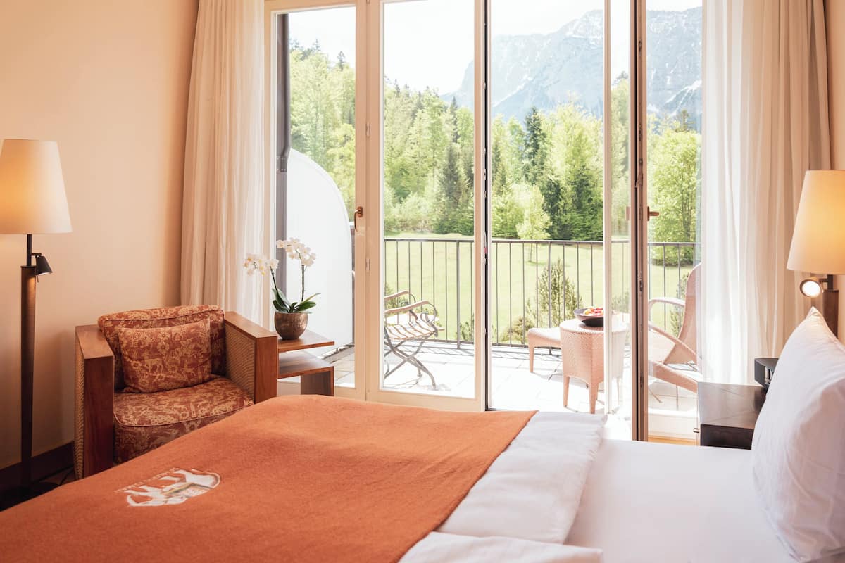 A hotel room with a bed, a chair, a table, and a balcony with a view of trees and mountains.