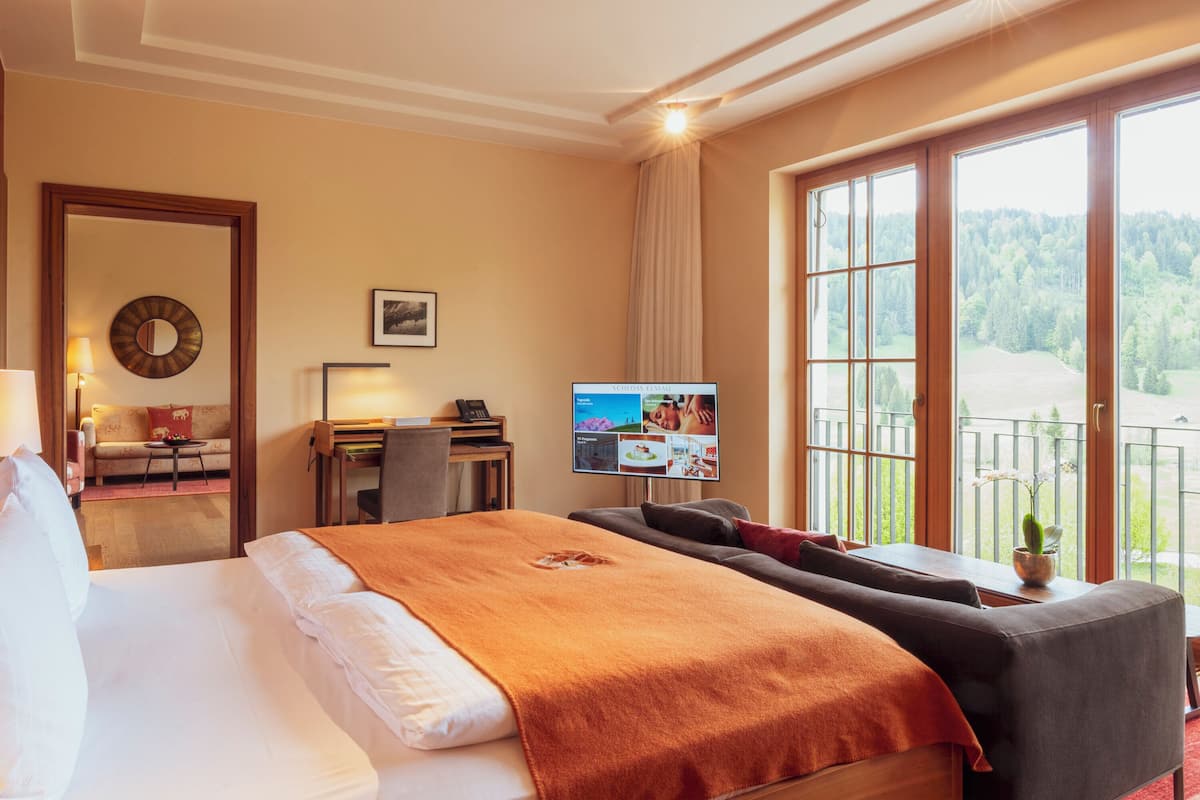 A hotel room with a large bed, a desk with a computer, a television, and a balcony with a view of the forest.
