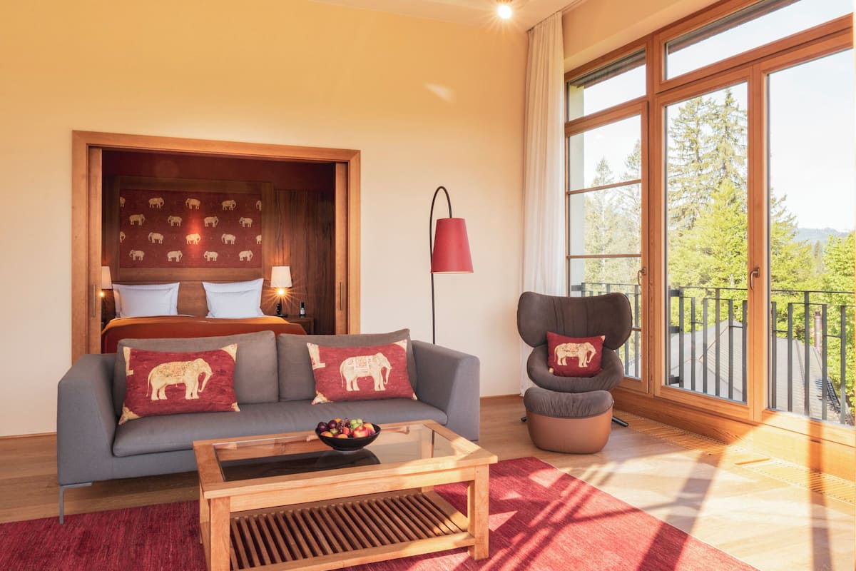 A modern living room with a sofa, a coffee table, and a chair. There is a large window with a view of trees and a balcony.
