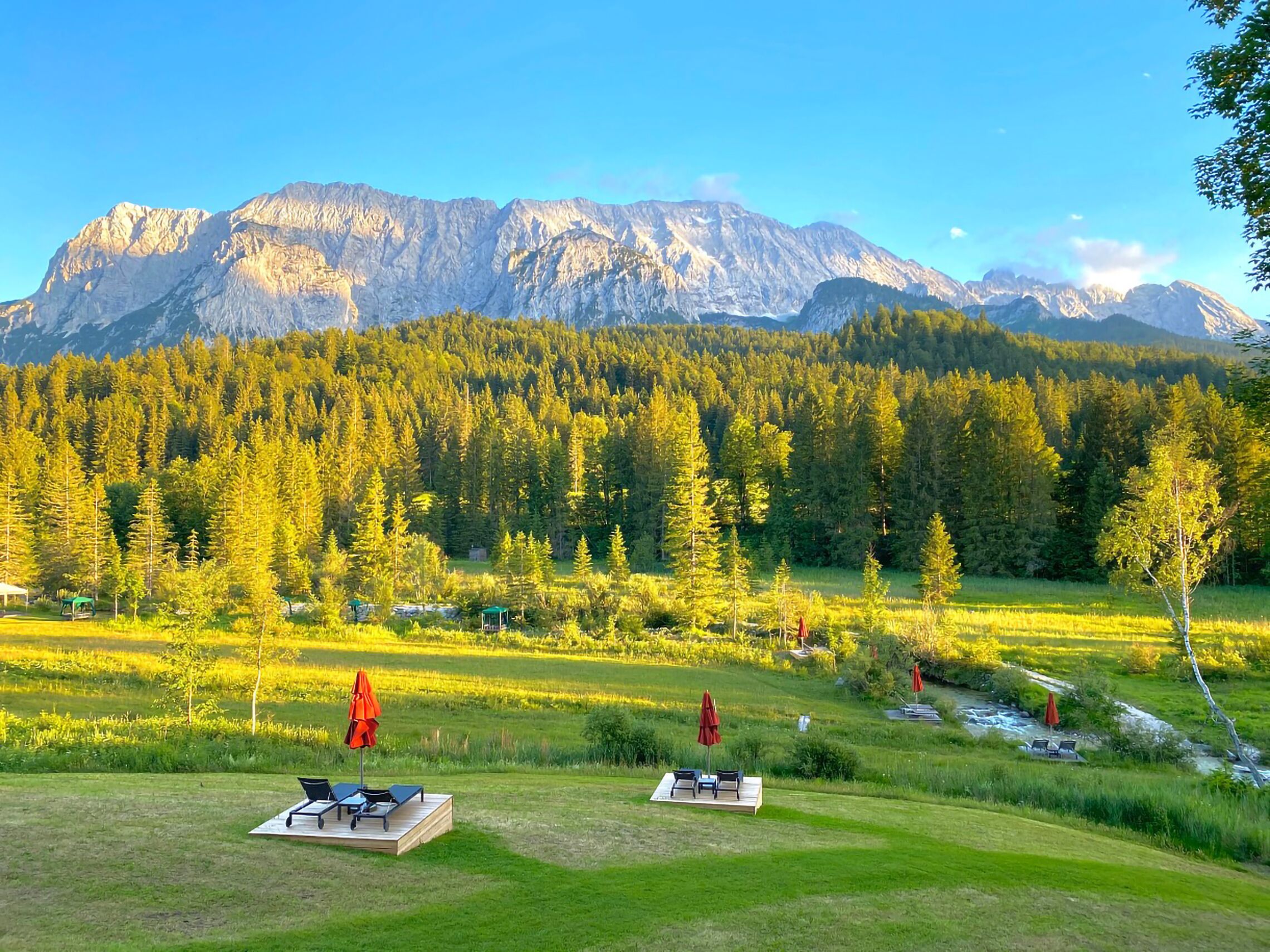 Eine friedliche Außenanlage mit Bergblick, mit einer Rasenfläche, zwei Liegestühlen und einer kleinen Terrasse, umgeben von dichtem Wald und blauem Himmel.
