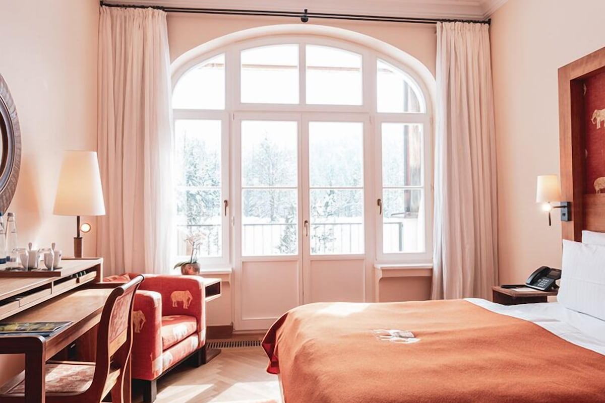 A hotel room with a large arched window, a bed with a brown cover, a desk with a lamp, and a chair.