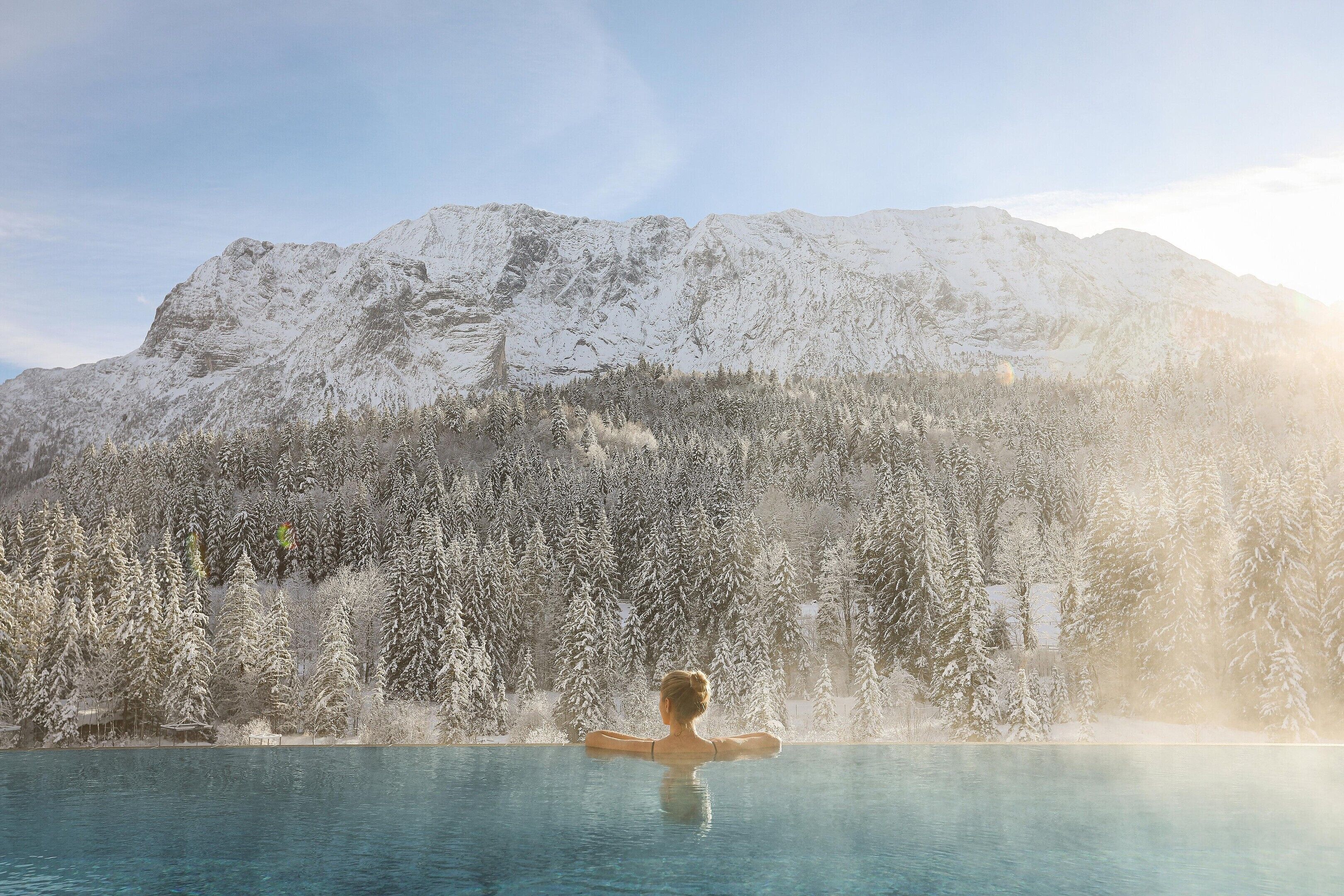 Eine Person entspannt in einem klaren Pool vor einer verschneiten Bergkulisse.