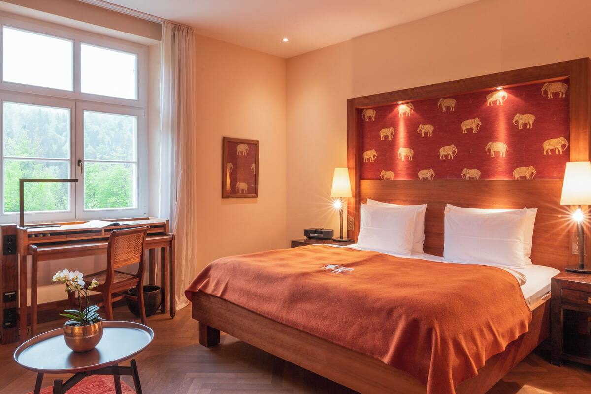 A hotel room with a large bed, two bedside tables with lamps, a desk with a chair, and a window with a view of greenery.