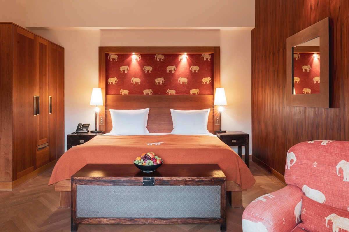 A hotel room with a large bed, wooden headboard, two bedside tables with lamps, a small table with fruit, and a red upholstered chair.