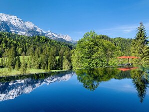 A serene lake with a clear reflection of trees and mountains, a red-roofed structure, and a wooden dock. - Schloss Elmau Luxury Spa Retreat & Cultural Hideaway (Kruen)