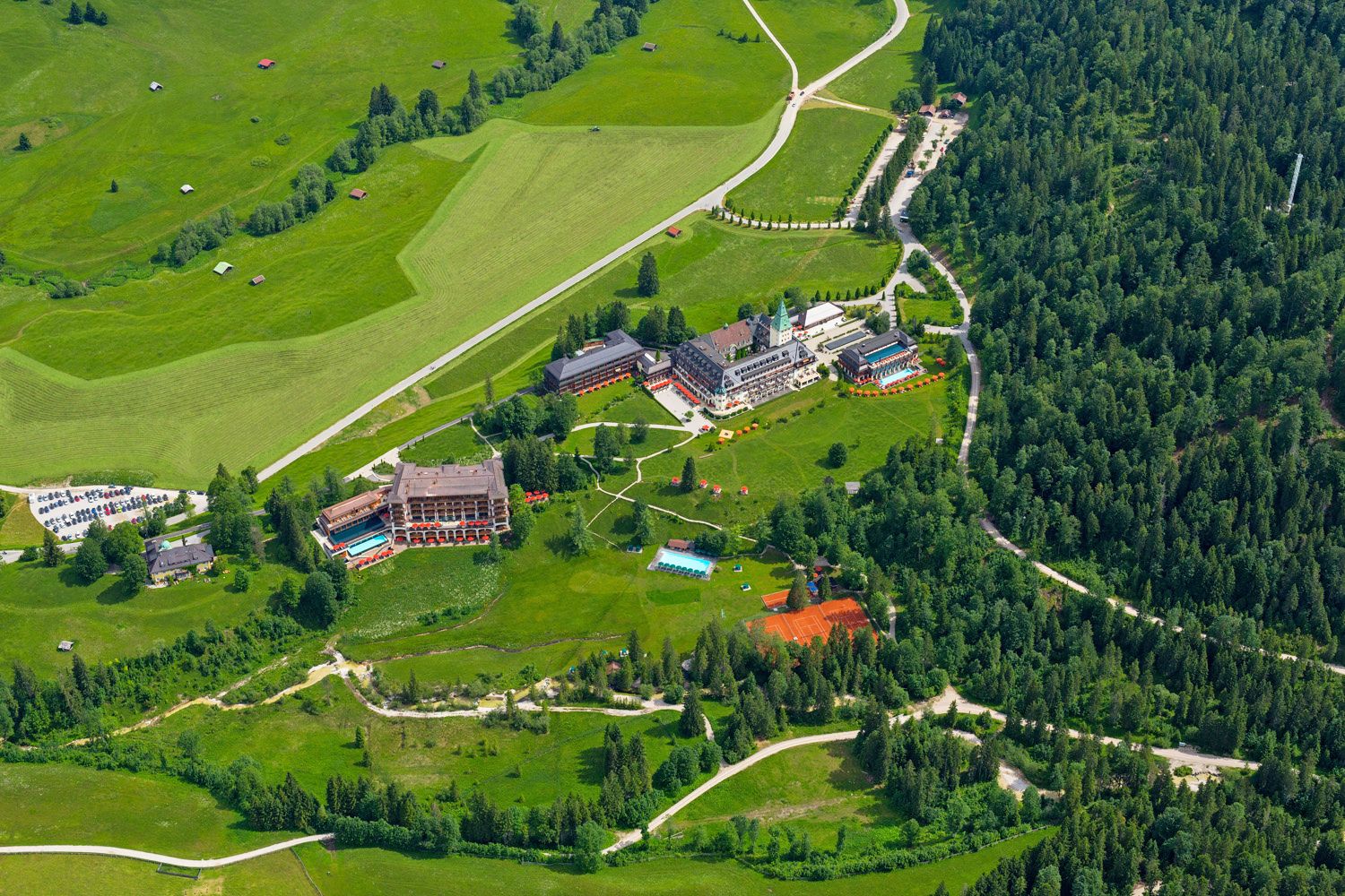 An aerial view of a resort with multiple buildings, including a large hotel with a swimming pool, surrounded by lush green fields and forested areas.