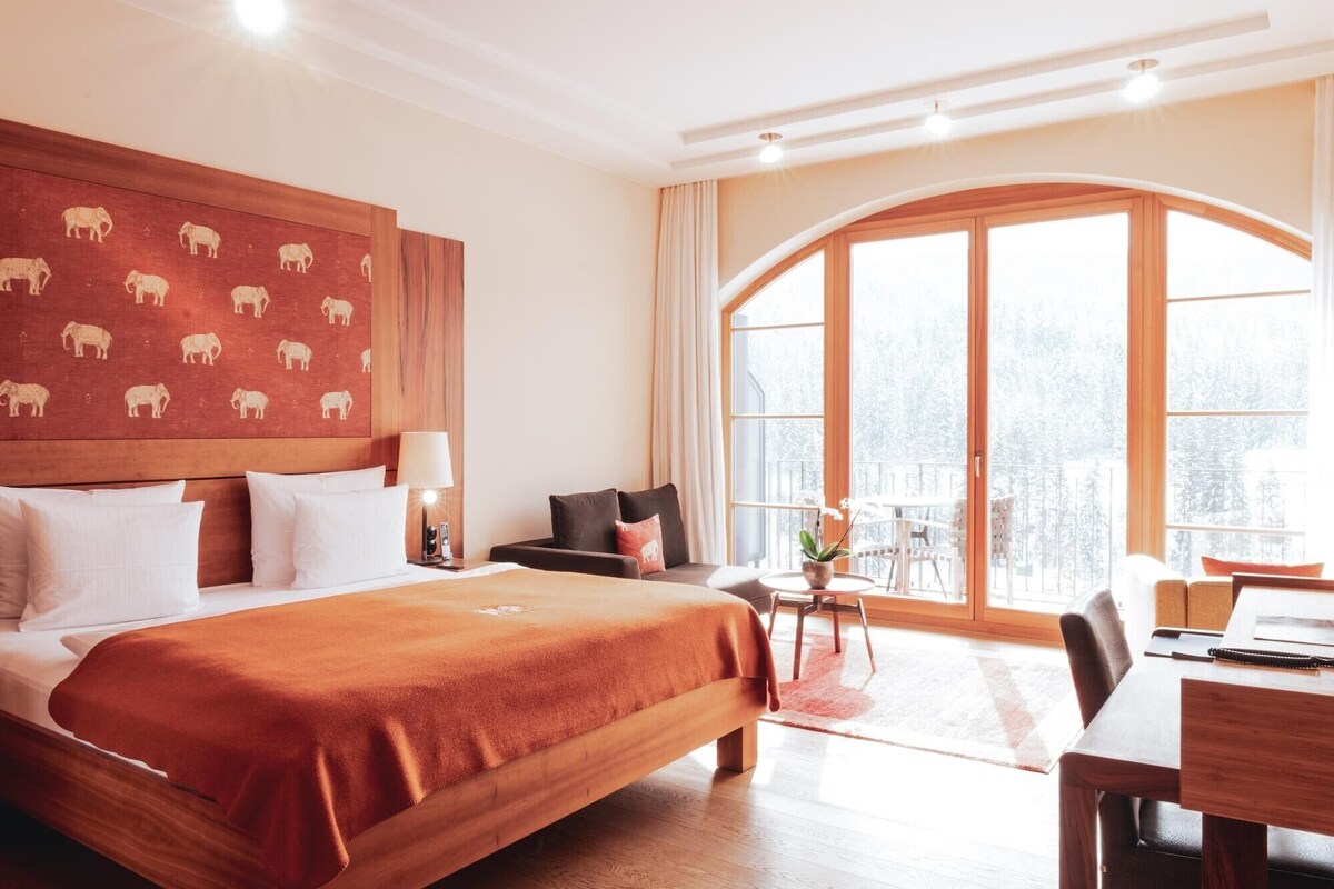 A hotel room with a large bed, a desk, a chair, a sofa, and a large window with a view.