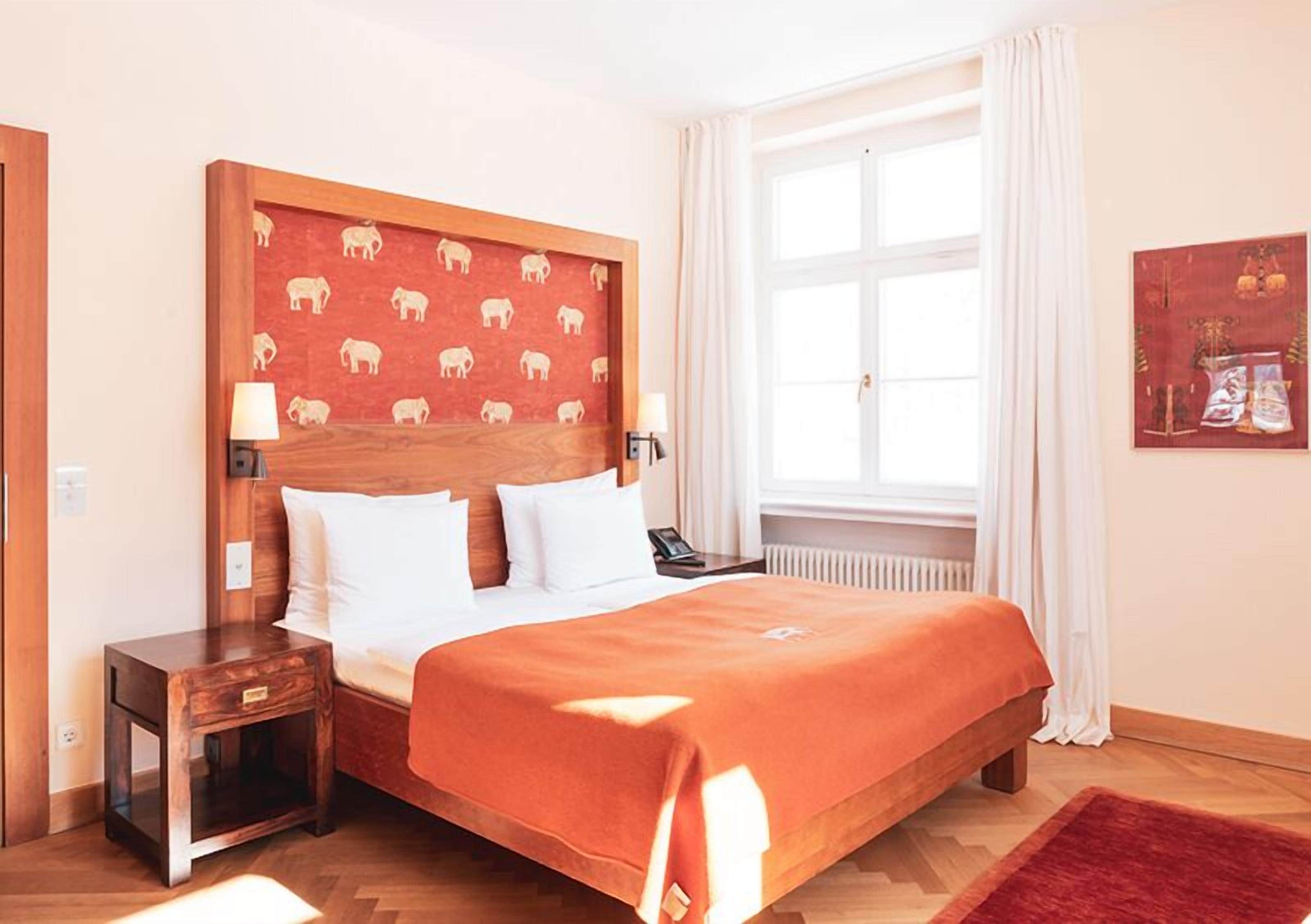 Een hotelkamer met een houten hoofdeinde, wit beddengoed, een rode plaid, een nachtkastje met een lamp en een raam met witte gordijnen.