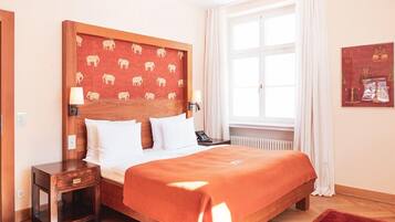 Een hotelkamer met een houten hoofdeinde, wit beddengoed, een rode plaid, een nachtkastje met een lamp en een raam met witte gordijnen.