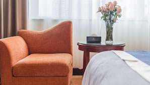Une chambre d’hôtel avec un seul fauteuil orange, une petite table ronde ornée d’un vase de fleurs roses et un lit parfaitement fait.