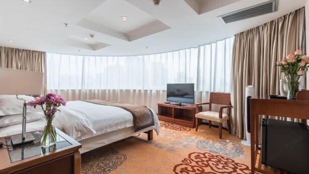 Une chambre d’hôtel avec un lit, une télévision, une chaise, un bureau avec une lampe et un vase de fleurs.