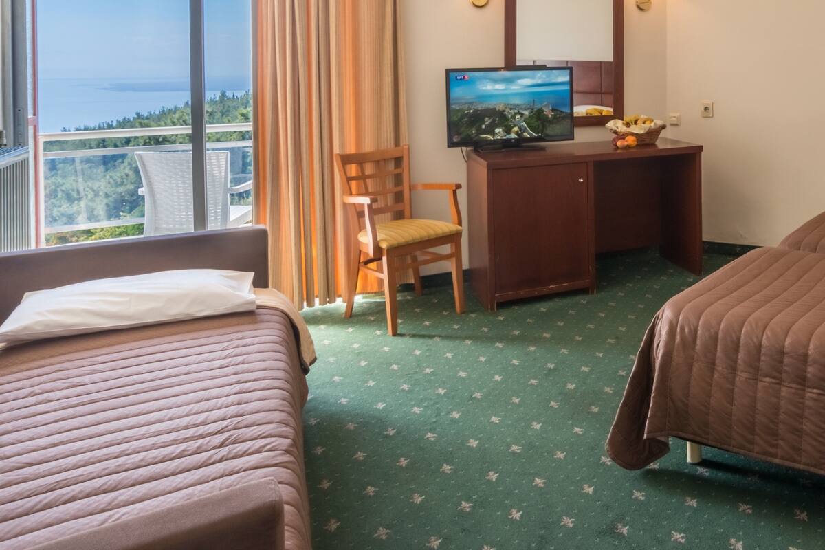 A hotel room with two beds, a desk with a television, a chair, and a view of the city and sea through large windows.