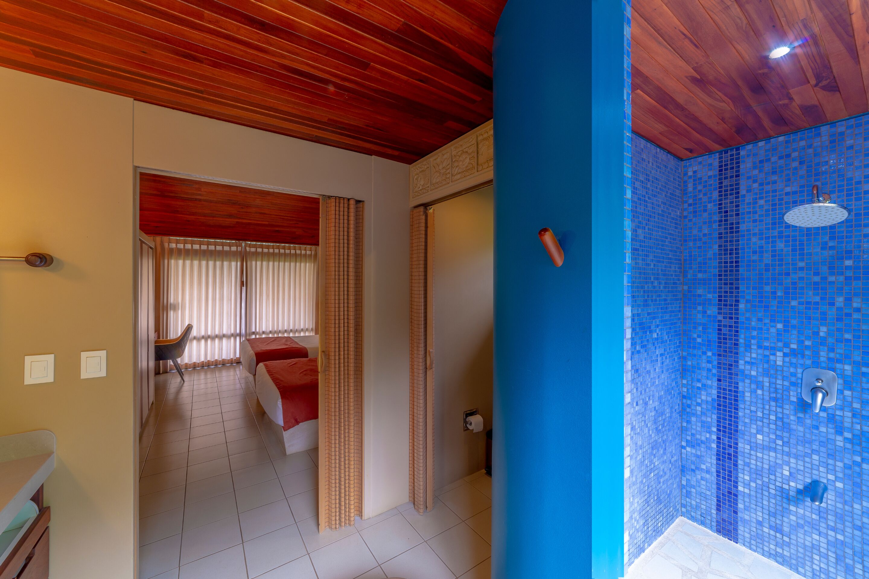 A hotel room with a wooden ceiling, a bathroom with blue mosaic tiles, and a bed with red bedding.