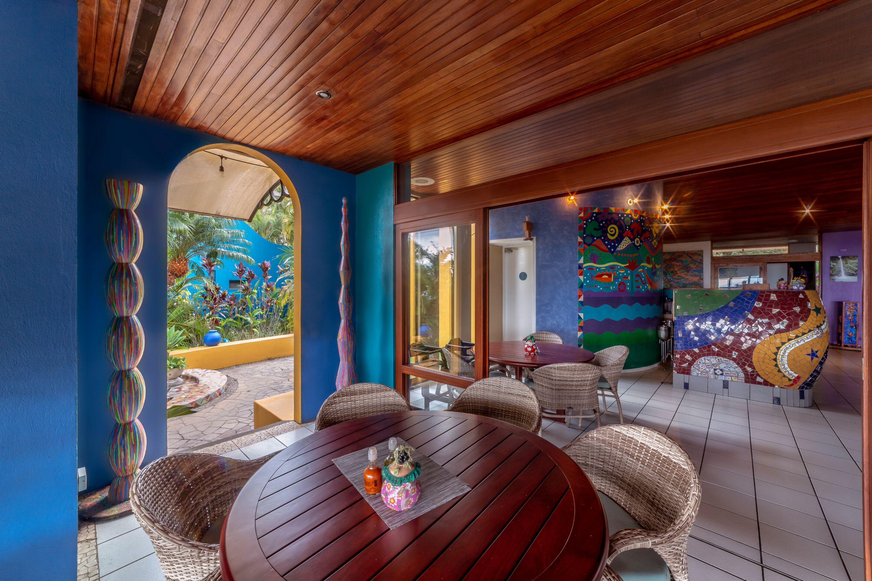 Un comedor con mesas redondas de madera y sillas de mimbre, un mural colorido en la pared, y vistas al exterior.