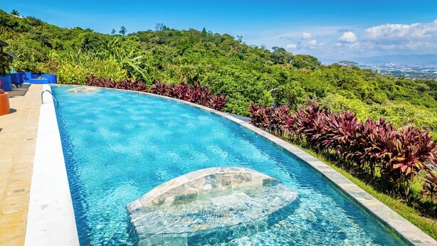 A swimming pool with a curved edge, surrounded by lush greenery and a view of a distant cityscape. - Xandari Resort And Spa (San Isidro)