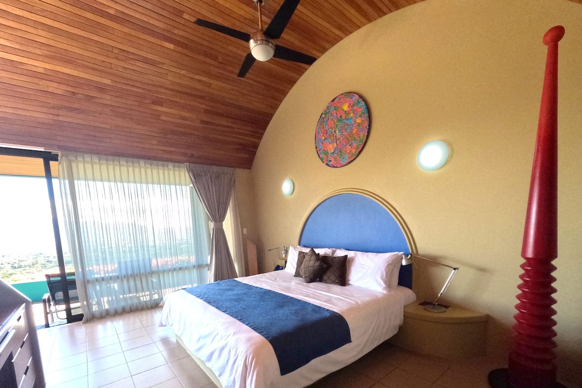 A bedroom with a bed, a ceiling fan, a nightstand, a lamp, and a view of the outside.