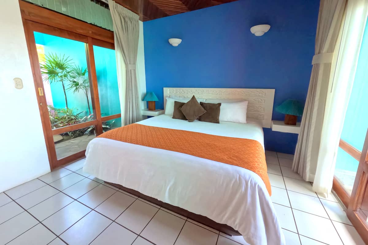 A hotel room with a bed, a nightstand, a window with curtains, and a view of palm trees.