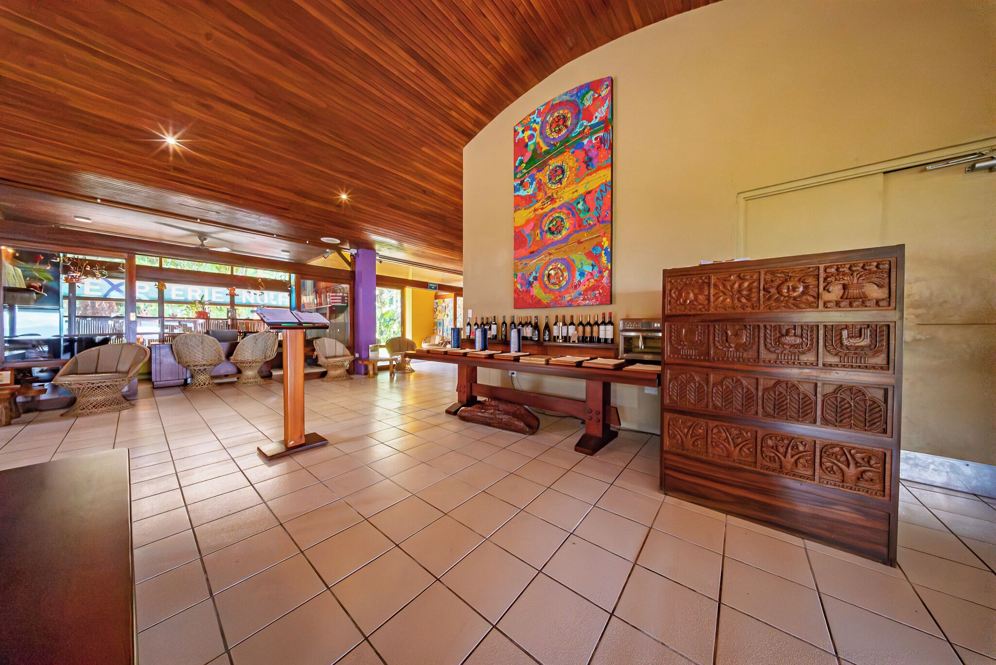 A spacious lobby with a wooden ceiling, a wine display, wicker furniture, and a colorful abstract painting.