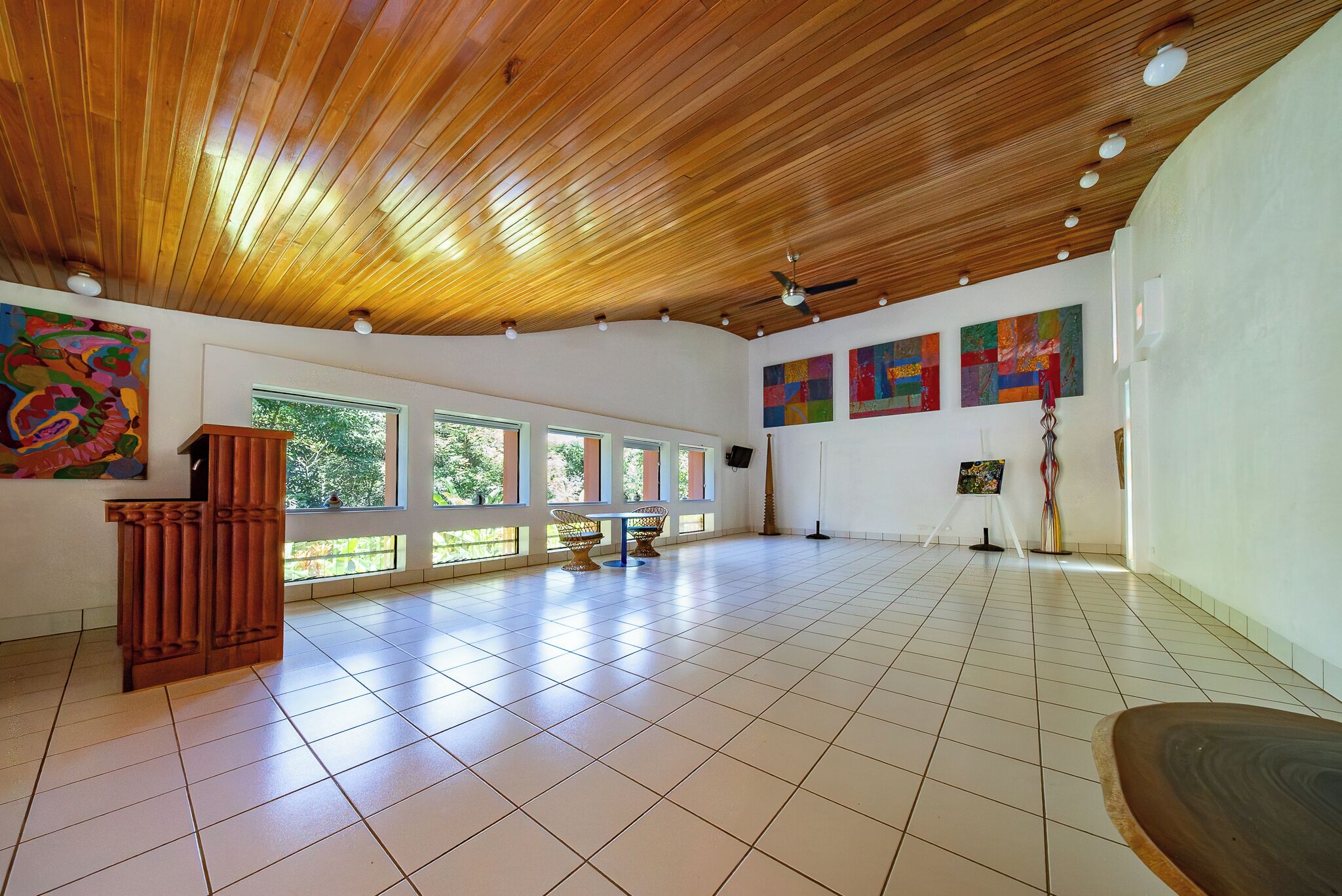 Una habitación amplia con techo de madera, piso de baldosas, ventanas grandes y obras de arte en las paredes.