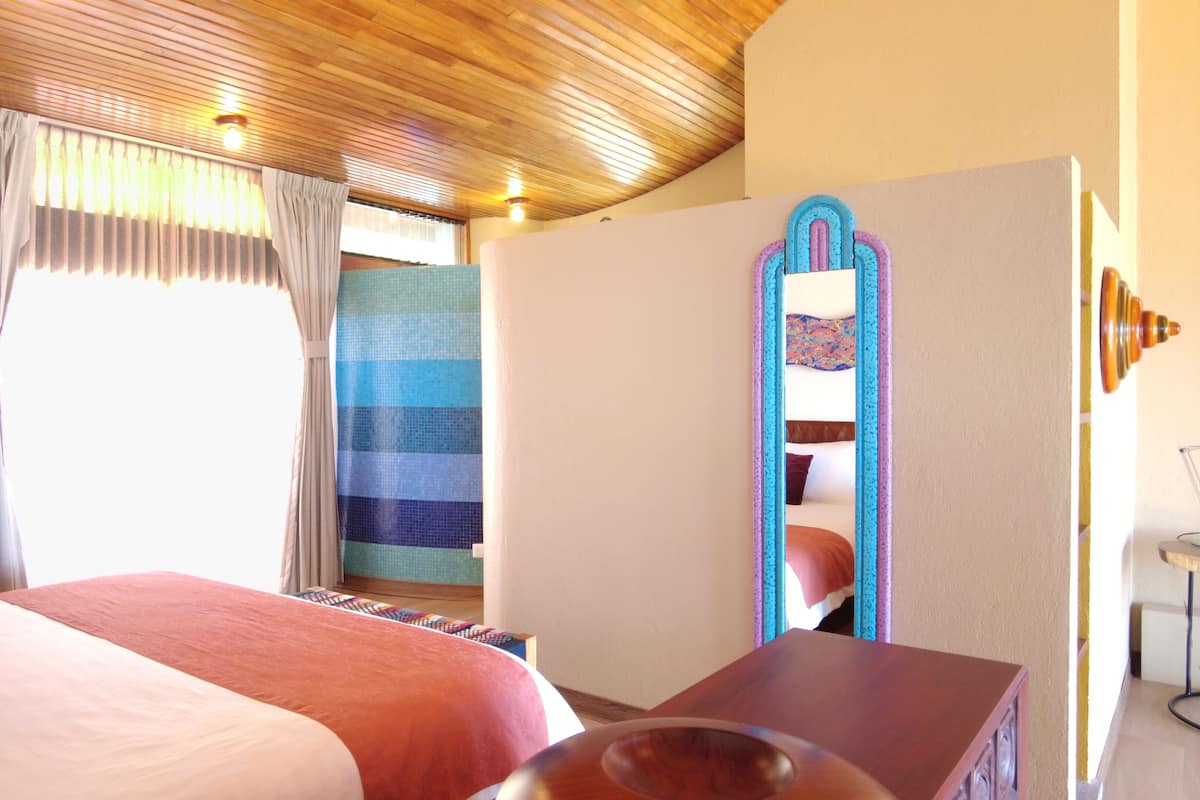 A hotel room with a wooden ceiling, a bed with a pink and white bedspread, a wooden nightstand, a mirror with a blue frame, and a window with curtains.