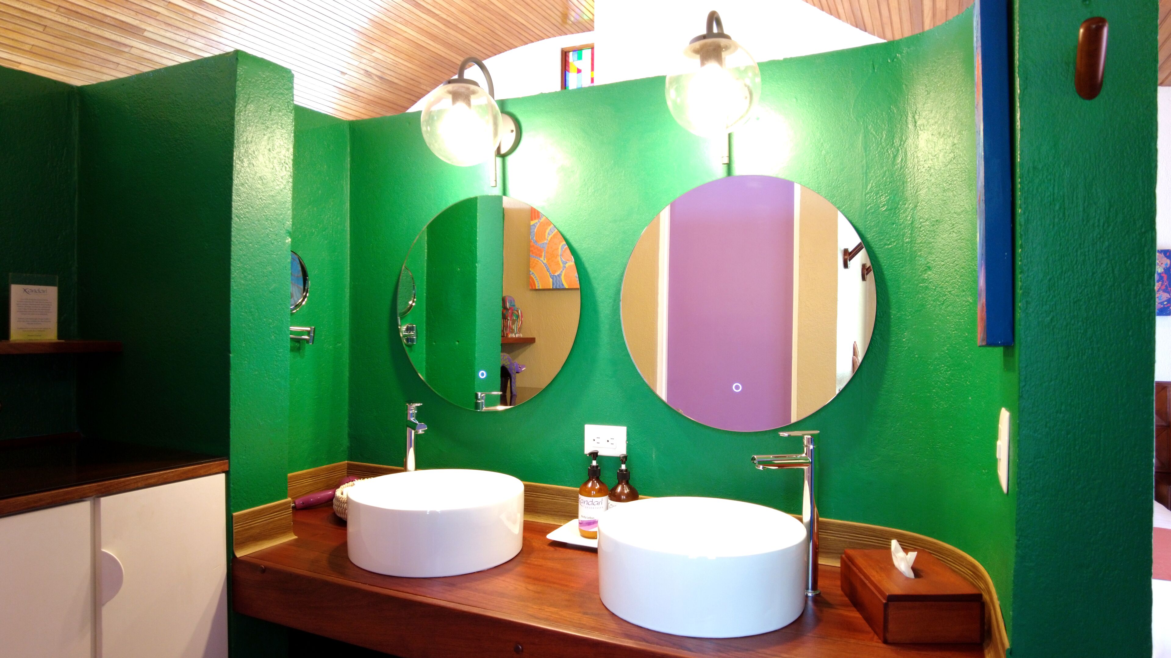 Une salle de bain aux murs verts, dotée de deux miroirs ronds et de plans de travail en bois.