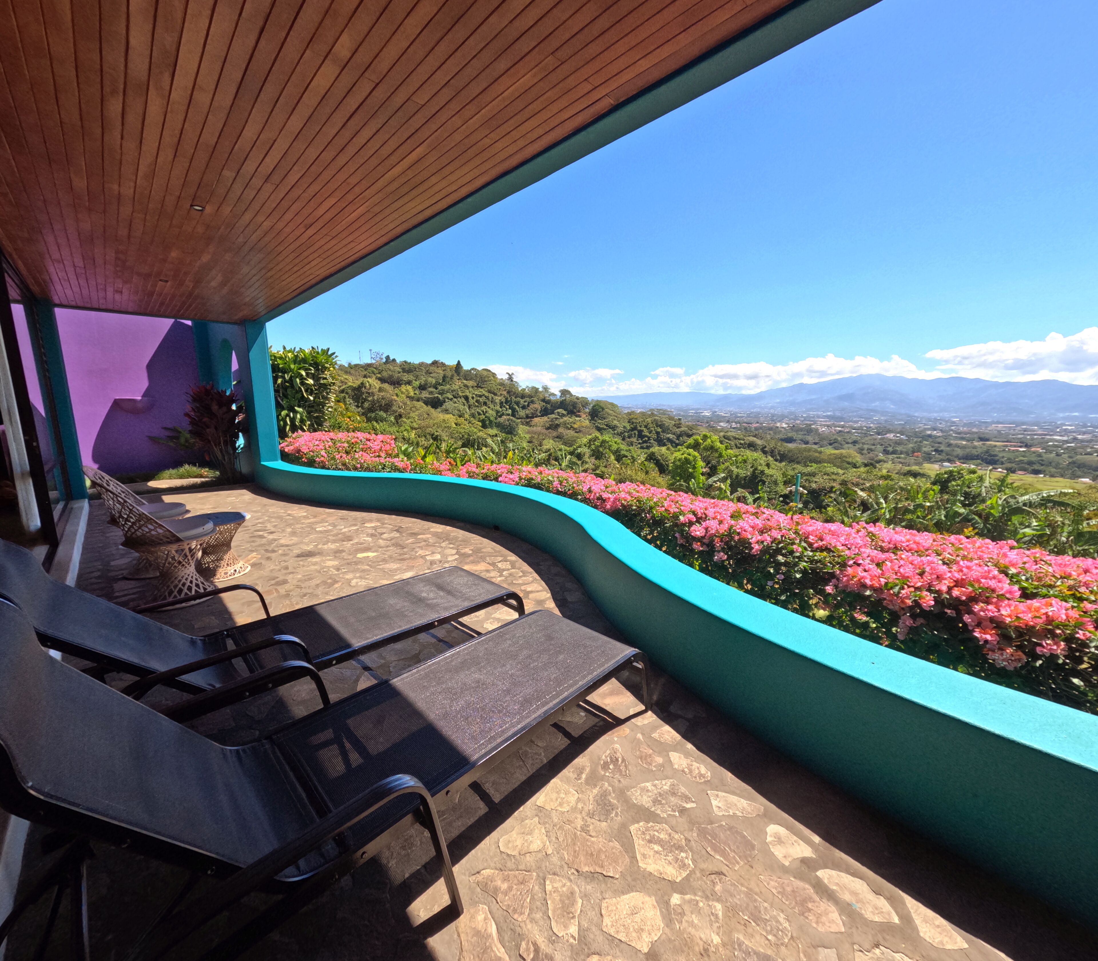 A covered outdoor seating area with lounge chairs, a view of a lush landscape with pink flowers, and distant mountains.