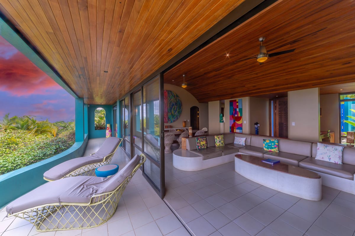 A modern lounge area with a curved seating arrangement, a ceiling fan, and large windows offering a view of the outdoors.
