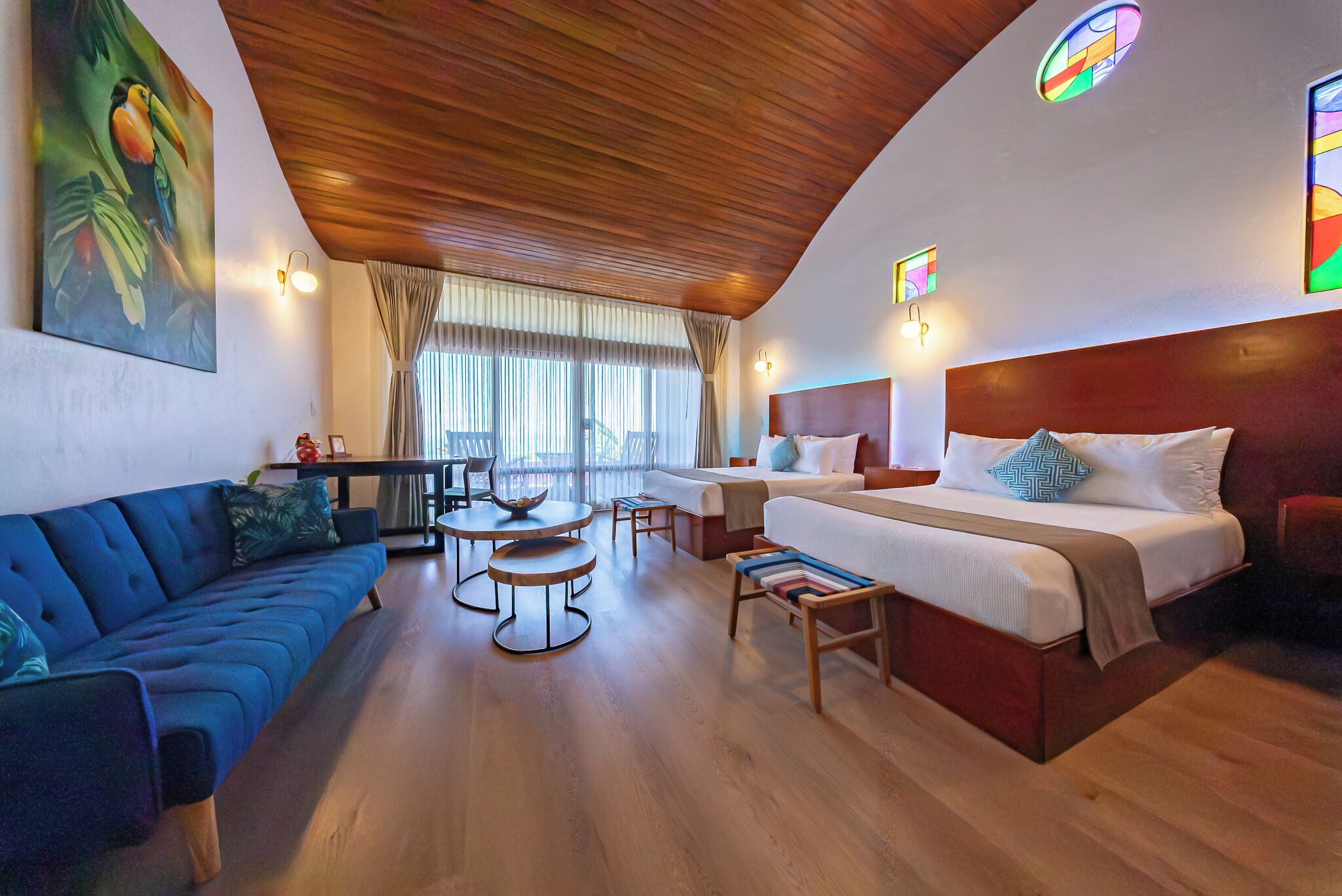 A hotel room with a wooden ceiling, a bed with a headboard, a sofa, a dining table, and a view of the outdoors.