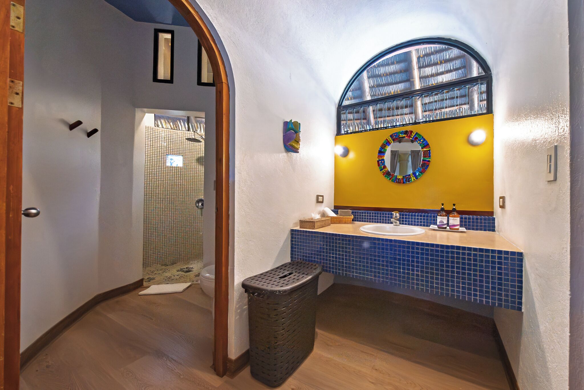 Une salle de bain avec un plancher en bois, un mur jaune, un lavabo dont le plan est recouvert de carreaux bleus, un miroir avec un cadre coloré et une douche dont le mur est également recouvert de carreaux.