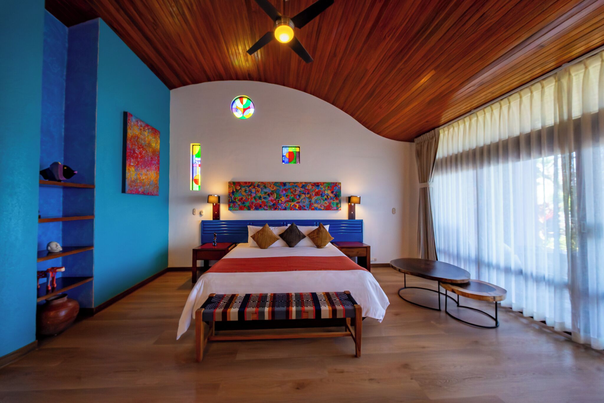 Une chambre à coucher avec un plafond incurvé, un lit doté d’une tête de lit colorée, un banc, une petite table et une fenêtre ornée de rideaux légers.