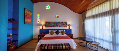 Une chambre à coucher avec un plafond incurvé, un lit doté d’une tête de lit colorée, un banc, une petite table et une fenêtre ornée de rideaux légers.