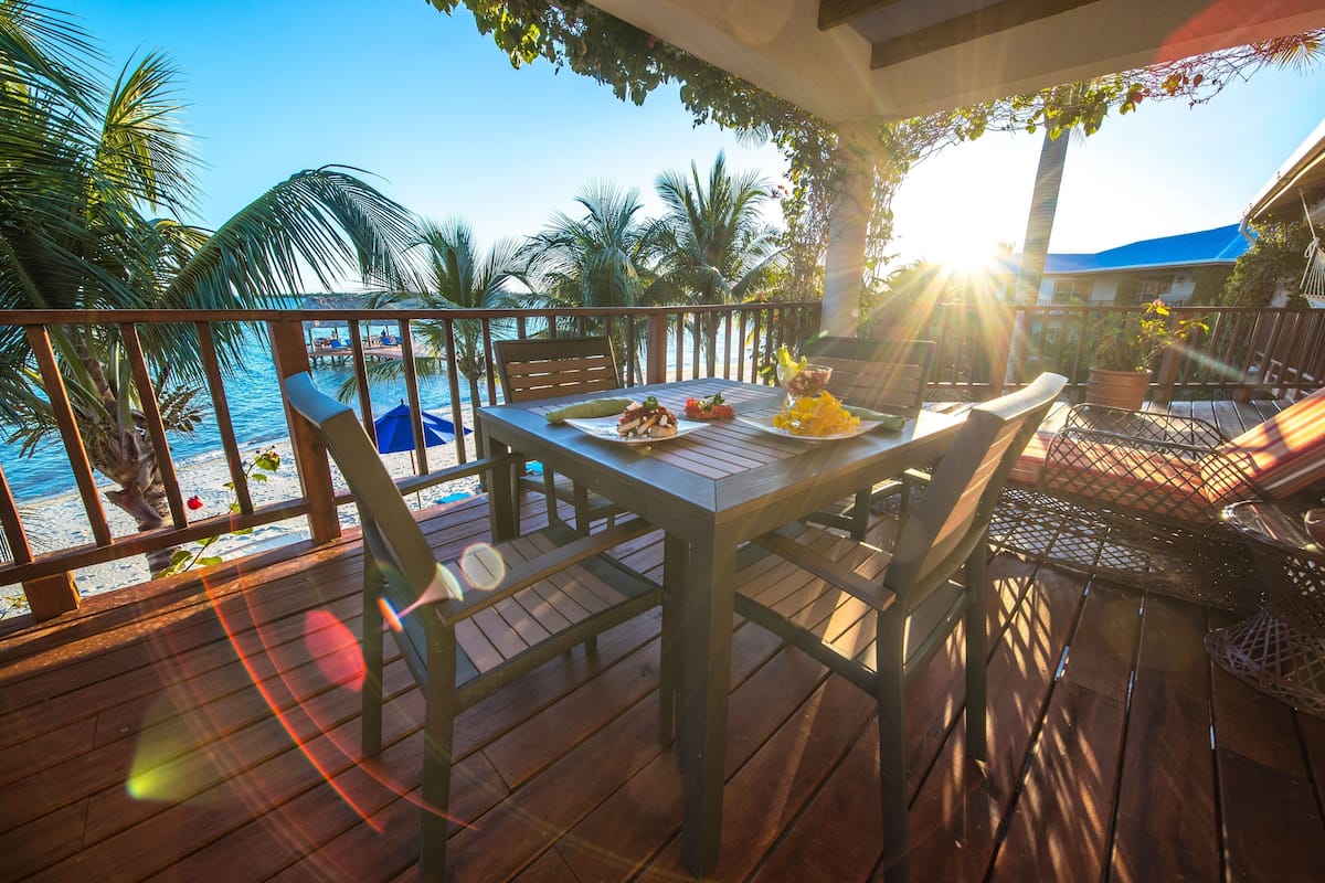 Eine Holzterrasse mit Tisch und Stühlen, mit Blick auf einen Strand mit Palmen und einem klaren Himmel.