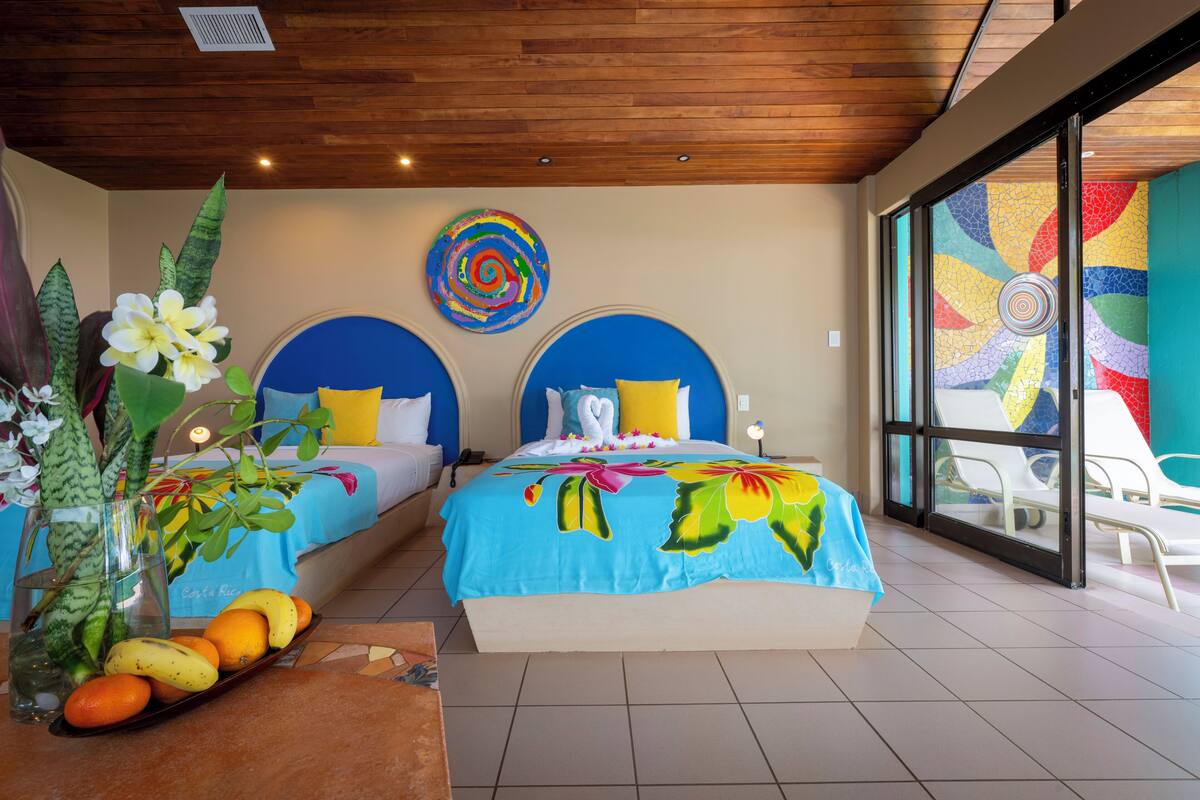 A hotel room with two beds, a large window with a view, and a tropical-themed bedspread.