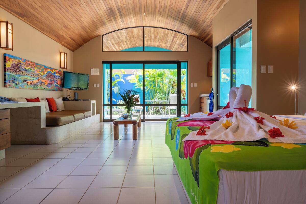 A spacious room with a large bed, a sofa, a television, and a view of palm trees through the glass doors.