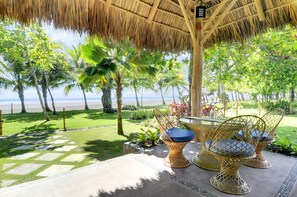 Eine Terrasse direkt am Strand mit Korbstühlen und einem Tisch, umgeben von Palmen und einem Reetdach.