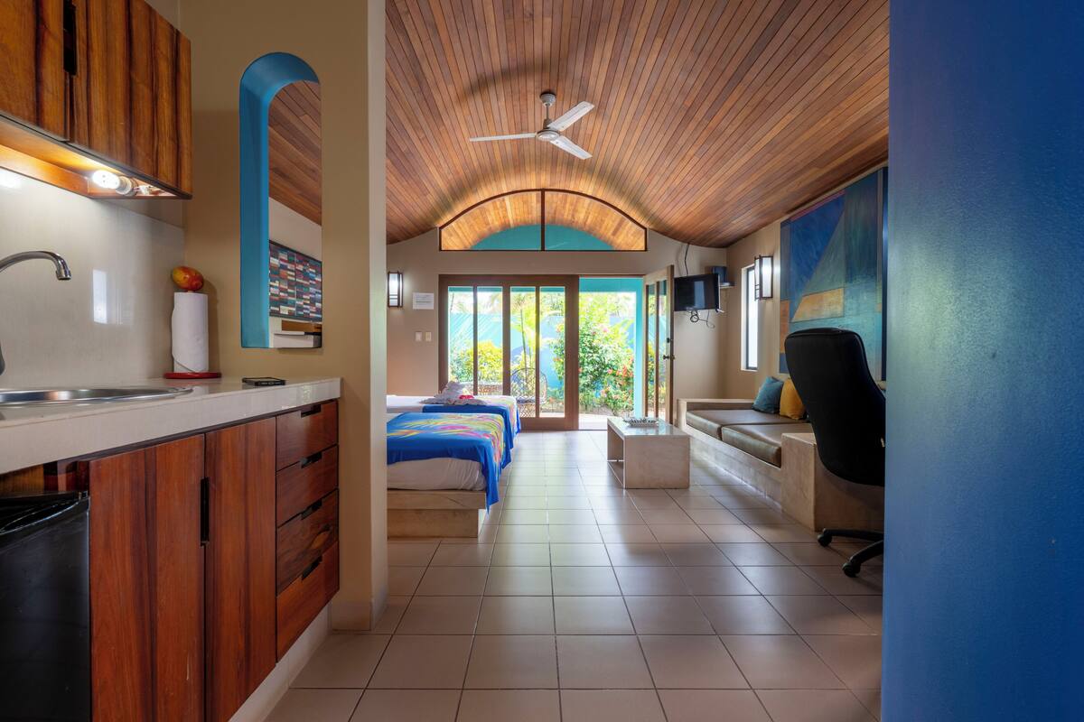 A modern hotel room with a kitchen, a bed with blue bedding, a desk, and a chair.