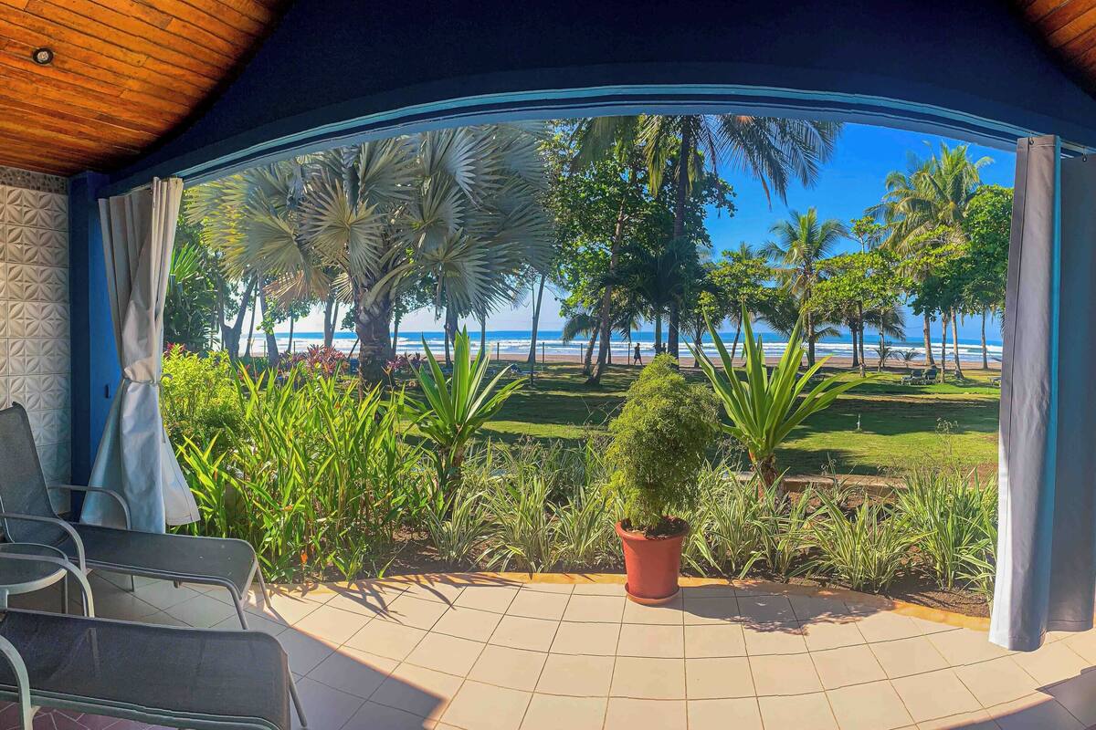 A covered patio with a view of the beach, outdoor seating, and potted plants.
